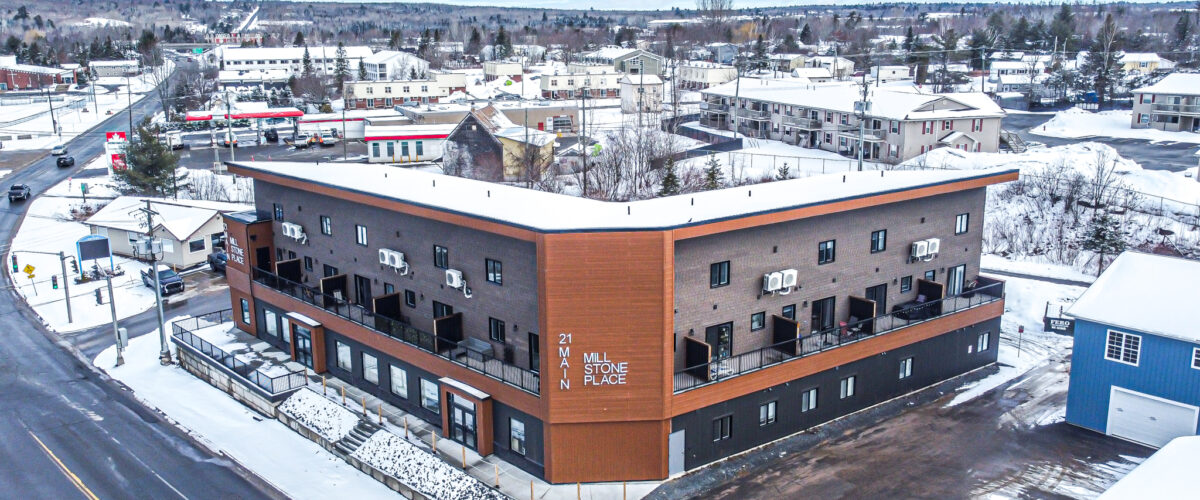 Aerial image of a grey and brown modern mixed use L-shaped building with ground level commercial space and two-level apartments above
